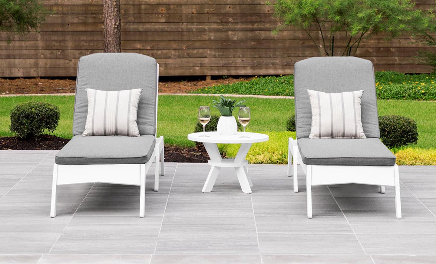 Two gray cushioned lounge chairs with striped pillows sit on a tiled patio, showcasing a minimalistic design. A Ledge Lounger Mainstay Round Side Table between them holds two glasses of white wine. The outdoor space is framed by lush bushes, trees, and a wooden fence.