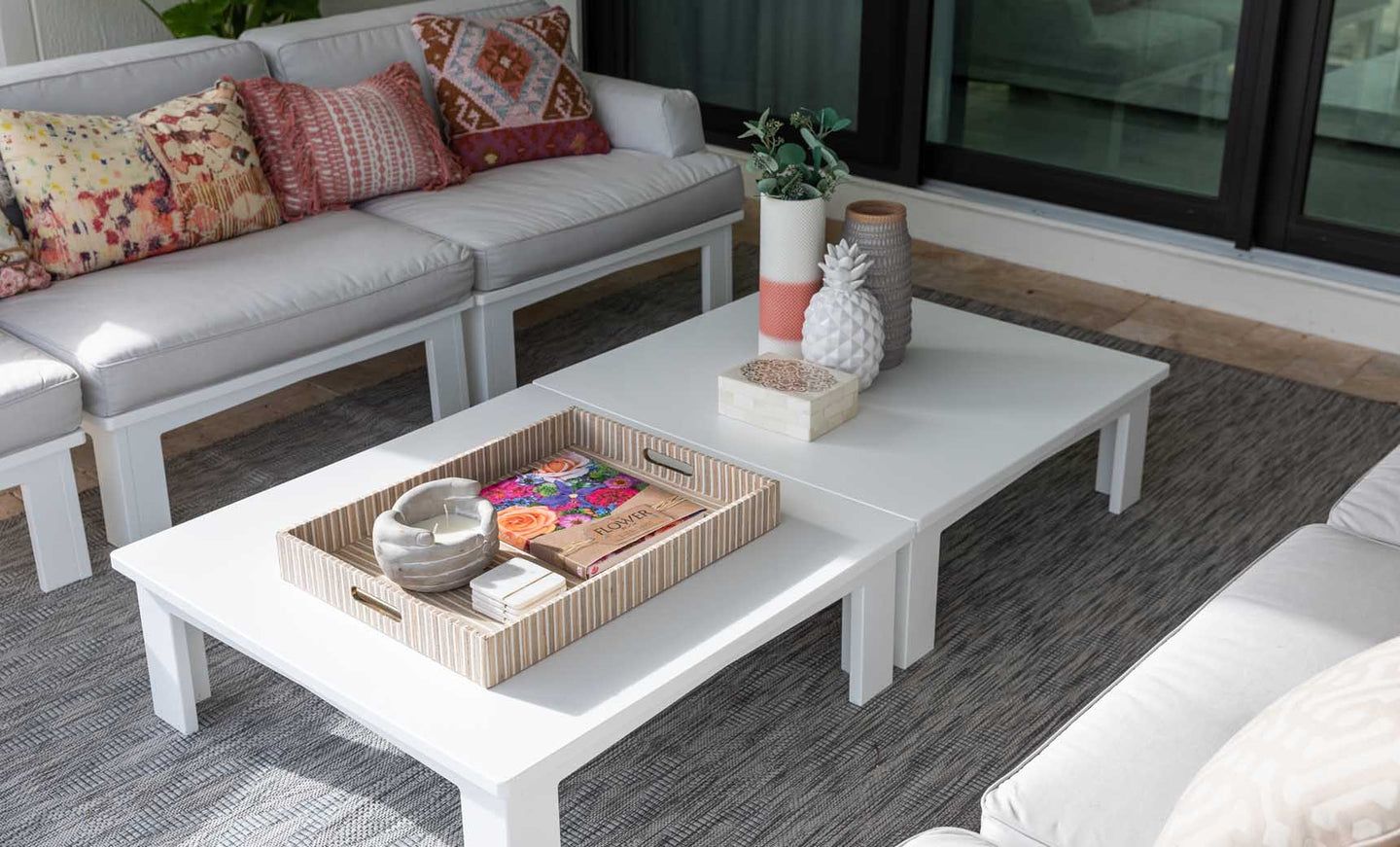 In a bright, modern living room, the Mainstay Square Coffee Table by Ledge Lounger is adorned with decorative items like a magazine tray, a pineapple ornament, and a plant. Surrounding it are white sofas from the same collection, accented with colorful patterned cushions.