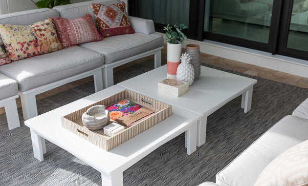 In a bright, modern living room, the Mainstay Square Coffee Table by Ledge Lounger is adorned with decorative items like a magazine tray, a pineapple ornament, and a plant. Surrounding it are white sofas from the same collection, accented with colorful patterned cushions.
