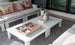 In a bright, modern living room, the Mainstay Square Coffee Table by Ledge Lounger is adorned with decorative items like a magazine tray, a pineapple ornament, and a plant. Surrounding it are white sofas from the same collection, accented with colorful patterned cushions.