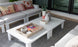 In a bright, modern living room, the Mainstay Square Coffee Table by Ledge Lounger is adorned with decorative items like a magazine tray, a pineapple ornament, and a plant. Surrounding it are white sofas from the same collection, accented with colorful patterned cushions.