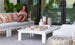 A woman lounges on a white outdoor sofa with colorful pillows, holding a drink and tablet. In front of her is the Mainstay Square Coffee Table by Ledge Lounger, showcasing decorative items as large glass doors open to a scenic view.