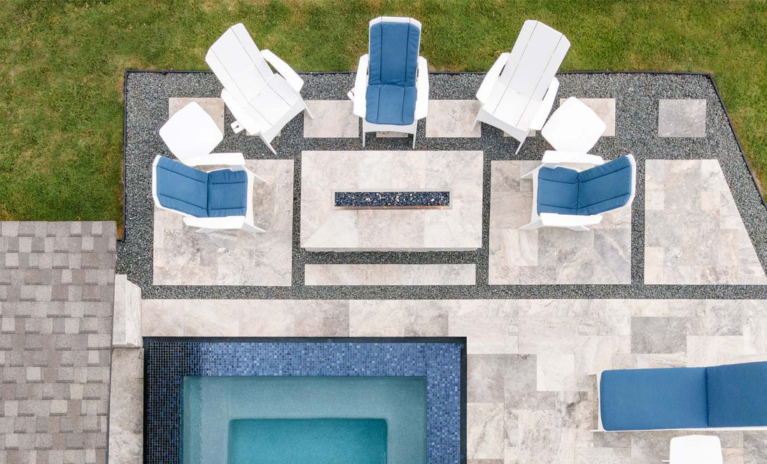 An aerial view of a modern patio shows six white chairs with blue cushions encircling a rectangular fire pit set on tile flooring with gravel accents, next to a small tiled pool. The scene includes the Mainstay Square Side Table by Ledge Lounger and nearby visible grass.