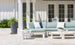 A patio features a light blue sectional sofa with white cushions, paired with a Mainstay Square Side Table in white holding decorative items. A tall planter with greenery sits on the left, and the background displays a white house with double glass doors.
