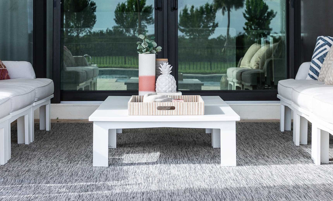 A modern outdoor seating area with white cushioned sofas, a Mainstay Square Coffee Table by Ledge Lounger on a gray rug, featuring a decorative tray with a vase and pineapple ornament. Glass sliding doors in the background reveal trees and a pool outside.