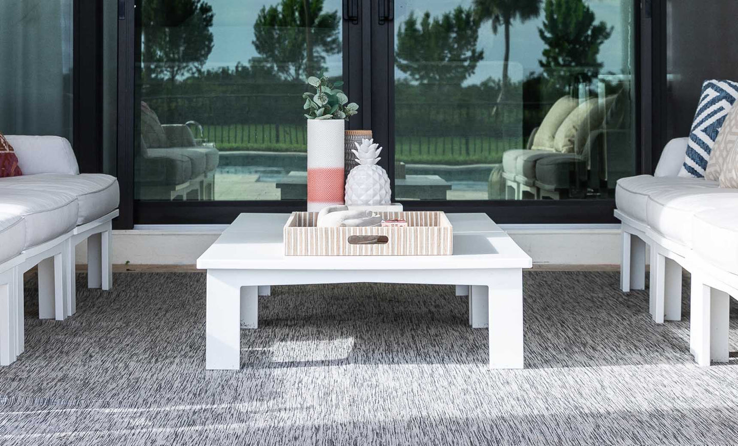 A modern outdoor seating area with white cushioned sofas, a Mainstay Square Coffee Table by Ledge Lounger on a gray rug, featuring a decorative tray with a vase and pineapple ornament. Glass sliding doors in the background reveal trees and a pool outside.