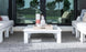 A modern outdoor seating area with white cushioned sofas, a Mainstay Square Coffee Table by Ledge Lounger on a gray rug, featuring a decorative tray with a vase and pineapple ornament. Glass sliding doors in the background reveal trees and a pool outside.