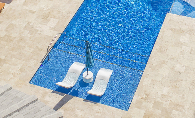 An aerial view showcases the Ledge Lounger Outdoor Oasis In-Pool Furniture Bundle: Signature Chaise Loungers partially submerged with an upright blue umbrella. The sparkling water glistens against beige stone tiles, creating a perfect outdoor escape.
