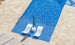 An aerial view showcases the Ledge Lounger Outdoor Oasis In-Pool Furniture Bundle: Signature Chaise Loungers partially submerged with an upright blue umbrella. The sparkling water glistens against beige stone tiles, creating a perfect outdoor escape.