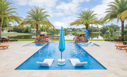 A luxurious outdoor pool showcases the Ledge Lounger Outdoor Oasis In-Pool Furniture Bundle, featuring two white chaise loungers partially submerged at one end. A blue umbrella stands between them for shade amid palm trees and a serene lake under a partly cloudy sky.
