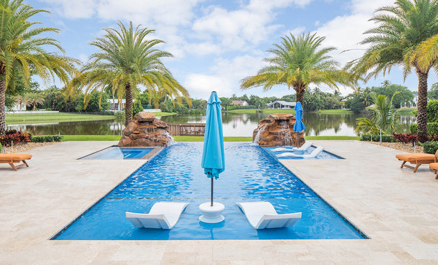 A luxurious outdoor pool showcases the Ledge Lounger Outdoor Oasis In-Pool Furniture Bundle, featuring two white chaise loungers partially submerged at one end. A blue umbrella stands between them for shade amid palm trees and a serene lake under a partly cloudy sky.