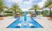 A luxurious outdoor pool showcases the Ledge Lounger Outdoor Oasis In-Pool Furniture Bundle, featuring two white chaise loungers partially submerged at one end. A blue umbrella stands between them for shade amid palm trees and a serene lake under a partly cloudy sky.