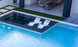 The modern pool features a raised rectangular section with Ledge Loungers Outdoor Paradise In-Pool Furniture Bundle, including sleek chaise loungers and a small table, surrounded by lush grass and a partially visible patio with inviting seating.