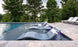 Ledge Loungers Outdoor Paradise In-Pool Furniture Bundle features luxurious chaise loungers with white cushions, set on stone tiles and surrounded by lush green trees, creating a serene atmosphere for ultimate relaxation.