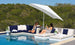 Two women relax and chat on modern poolside furniture, featuring Ledge Loungers Autograph Standard Side Table, cushioned loungers, and a sectional sofa under a large white umbrella, with trees and blue sky in the background.