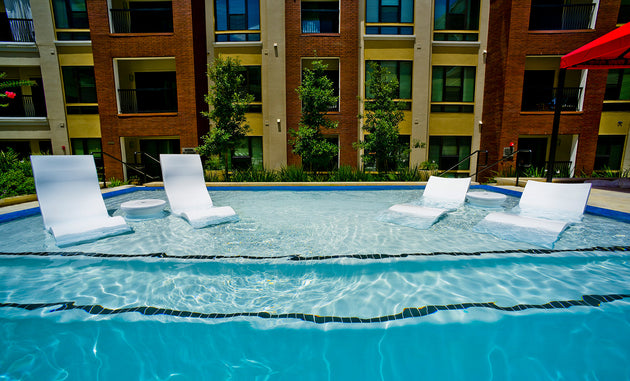 The Ledge Lounger Signature Chaise with Chairs and Side Table In-Pool 6-Piece Set sits partially submerged in a clear blue pool, with a modern apartment building and green trees visible in the background under bright sunlight.