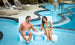 A man and woman relax on Ledge Lounger’s Signature Chaise with Chairs and Side Table In-Pool 6-Piece Set, smiling with drinks. Others gather by the pool steps, with sun loungers and lush plants lining the deck in the background.