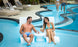 A man and woman relax on Ledge Lounger’s Signature Chaise with Chairs and Side Table In-Pool 6-Piece Set, smiling with drinks. Others gather by the pool steps, with sun loungers and lush plants lining the deck in the background.