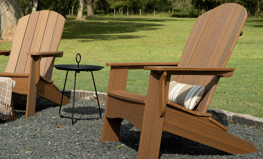 Two wooden Adirondack chairs with a Ledge Lounger 10x18 rectangular throw pillow rest on a pebble-covered area in a grassy yard. A small black metal table stands between them, with a plaid blanket draped over one arm of the chair. Trees and an expansive lawn are visible in the background.