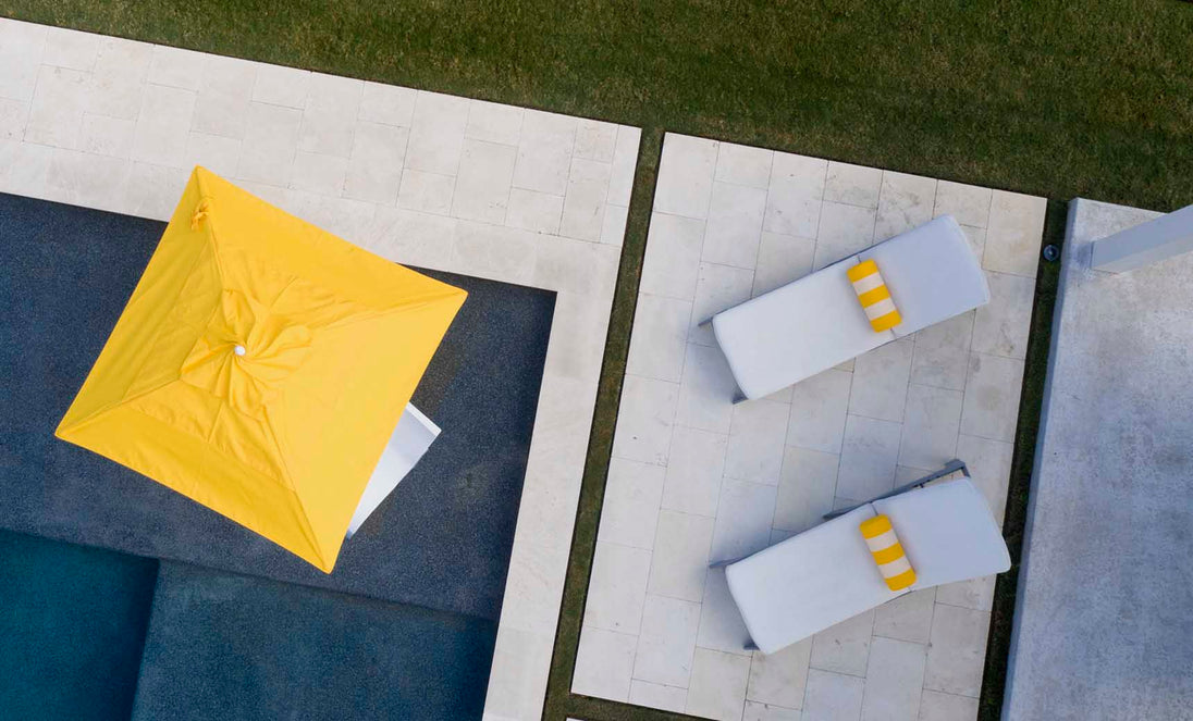 Aerial view of two white lounge chairs with EZ-Dri foam and Ledge Loungers Round Bolster Throw Pillows in yellow on a stone patio by a pool. A large yellow umbrella offers shade, vibrant outdoor fabrics are highlighted, and a green grass strip borders the patio.