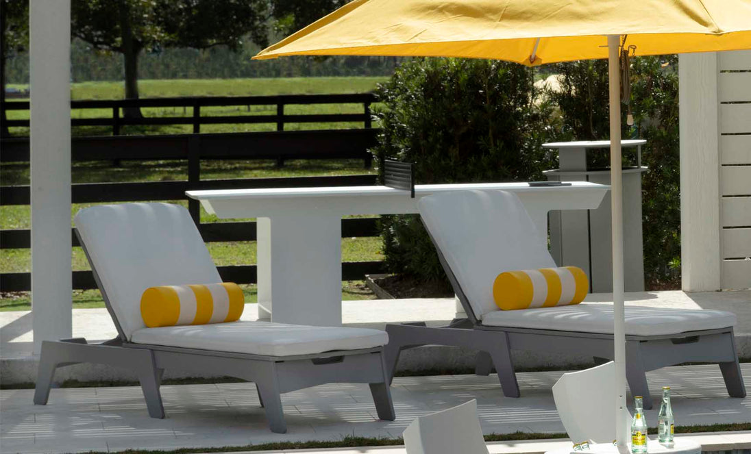Two Ledge Lounger recliners with white cushions and Round Bolster Throw Pillows in a yellow stripe pattern rest under a yellow umbrella. A nearby white table beautifully accents the surrounding greenery and black fence.