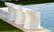 Three Ledge Lounger Signature Barstool Counter Height chairs with curved backs are lined up by a pool on a stone patio, bordered by trimmed grass. The sleek, modern chairs complement the glistening blue pool water in the sunlight.