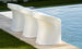 Three Ledge Lounger Signature Barstool Counter Height chairs with curved backs are lined up by a pool on a stone patio, bordered by trimmed grass. The sleek, modern chairs complement the glistening blue pool water in the sunlight.