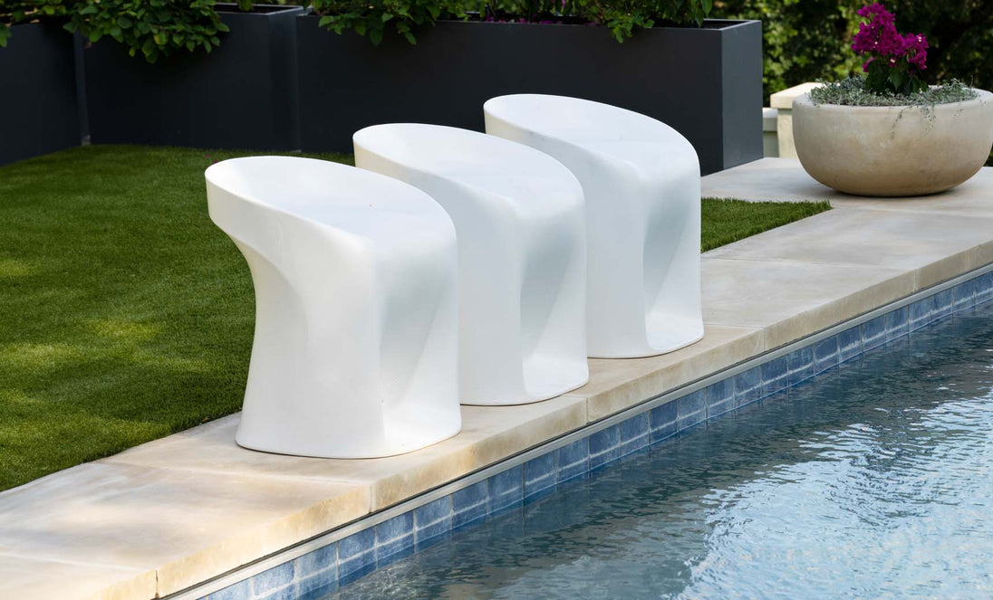 Three sleek Signature Barstool Counter Height stools by Ledge Lounger line a pools edge on a stone patio, with green plants in large pots and a neatly trimmed lawn in the background.