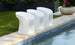 Three sleek Signature Barstool Counter Height stools by Ledge Lounger line a pools edge on a stone patio, with green plants in large pots and a neatly trimmed lawn in the background.