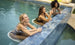 Three women enjoy drinks on Ledge Loungers Signature Barstool Bar Height at the pools edge, surrounded by vibrant greenery and tiled borders, as their laughter epitomizes outdoor entertaining.