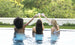 Three women in swimsuits relax at a pool, toasting with drinks. Surrounded by greenery and a white wall, they embody outdoor entertaining, exuding celebration and comfort from their perch on Ledge Loungers Signature Barstool Bar Height.