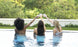 Three women in swimsuits relax at a pool, toasting with drinks. Surrounded by greenery and a white wall, they embody outdoor entertaining, exuding celebration and comfort from their perch on Ledge Loungers Signature Barstool Bar Height.