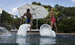 Two shirtless men wearing sunglasses and blue-red swim trunks playfully push white Signature Barstool Bar Height chairs by Ledge Lounger into the pool. An open white umbrella and lush green trees create an ideal outdoor entertaining backdrop.