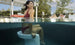 A woman in a red swimsuit sits on a submerged Ledge Lounger Signature Barstool Bar Height in the pool, smiling at the sky and capturing outdoor enjoyment. Nearby, someone relaxes by the pool under a pergola framed by trees.