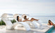 Three people in swimwear enjoy the sun on modern, white Signature Chaise loungers by Ledge Lounger overlooking a calm sea. Two recline with drinks while another kneels at the pools edge, drink in hand, all captured through a vivid media viewing window in this serene setting.