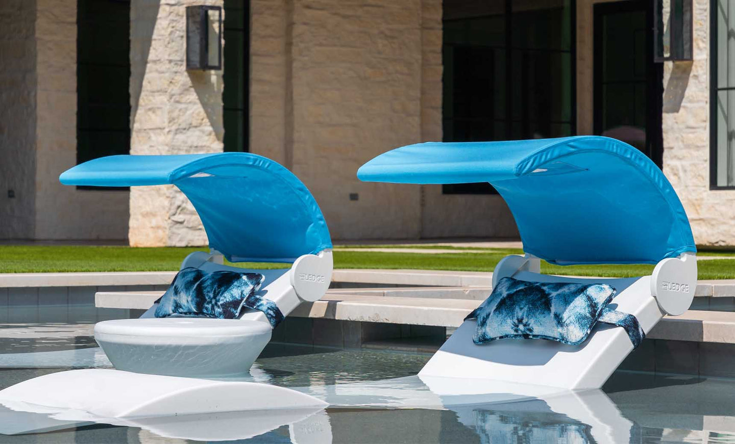 Two Ledge Lounger Signature Chaise Shade lounge chairs with blue canopies and patterned pillows sit in shallow pool water, perfect for sun relaxation. A stone building and green grass are visible in the background.