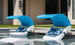 Two Ledge Lounger Signature Chaise Shade lounge chairs with blue canopies and patterned pillows sit in shallow pool water, perfect for sun relaxation. A stone building and green grass are visible in the background.