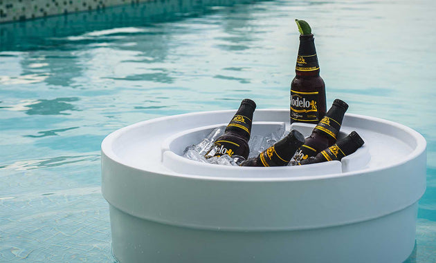 Five bottles of Modelo beer chill in a Signature Ice Basin by Ledge Lounger, filled with ice and floating on a clear blue swimming pool. One bottle stands upright with a lime wedge on the rim, while sunlight reflects off the water, creating a relaxing summer ambiance.
