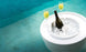 Floating in a pool, the Ledge Lounger Signature Ice Basin elegantly holds a bottle of champagne. Two filled glasses beside it create a luxurious, relaxing ambiance on the calm water.