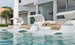 Three white lounge chairs are submerged in a modern pool outside a contemporary home with large glass doors. A small table, featuring the Signature Ice Basin by Ledge Lounger with a bottle and glasses, enhances the serene setting. Palm trees and a chaise lounge complete the scene.