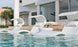 Three white lounge chairs are submerged in a modern pool outside a contemporary home with large glass doors. A small table, featuring the Signature Ice Basin by Ledge Lounger with a bottle and glasses, enhances the serene setting. Palm trees and a chaise lounge complete the scene.