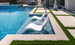 Two Ledge Lounger Signature Lowback Chairs are partially submerged in a modern pool, forming a serene outdoor oasis. A stone patio with artificial grass borders the area, while sunlight dances on the clear blue water beside a large decorative vase.
