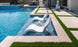 Two Ledge Lounger Signature Lowback Chairs are partially submerged in a modern pool, forming a serene outdoor oasis. A stone patio with artificial grass borders the area, while sunlight dances on the clear blue water beside a large decorative vase.