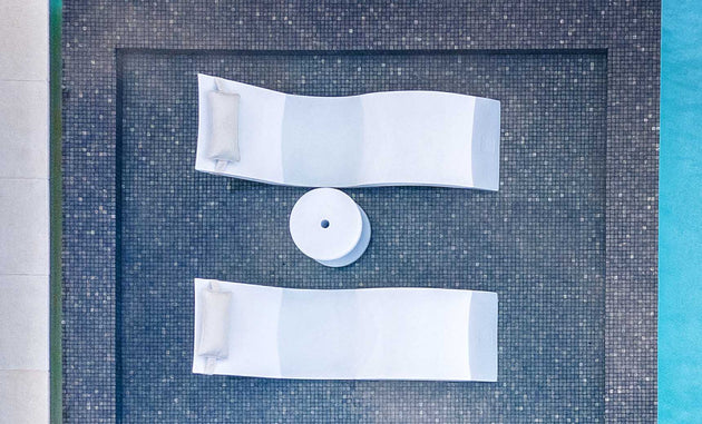 An aerial shot shows two white sun loungers with cushions on dark tiles by a pool. A Ledge Lounger Signature Standard Side Table, perfect for in-pool use or on the sun shelf, is placed elegantly between them.