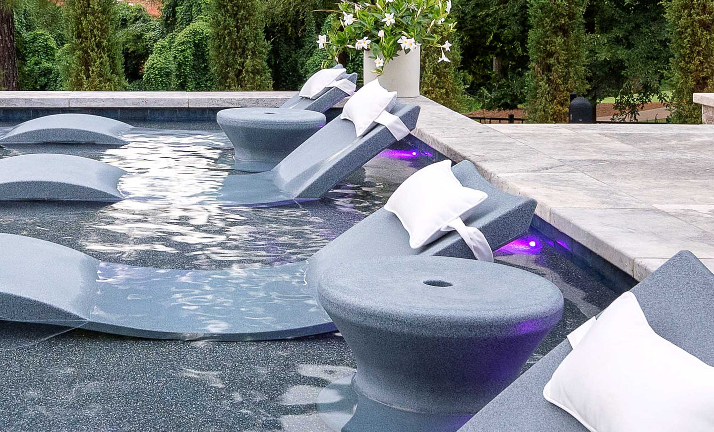 Submerged lounge chairs with white cushions and cup holders rest on a sun shelf under purple lights, perfect for in-pool use. The scene is enhanced by Ledge Loungers Signature Standard Side Table and a white flower arrangement among lush greenery.