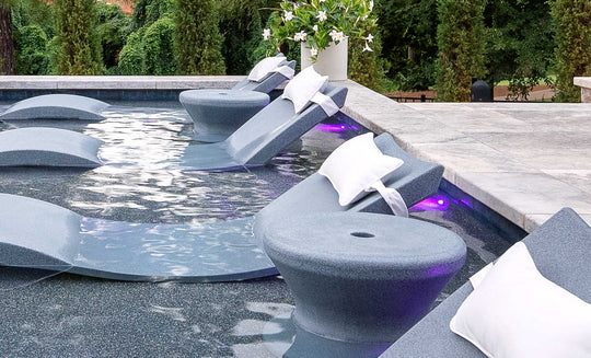 Submerged lounge chairs with white cushions and cup holders rest on a sun shelf under purple lights, perfect for in-pool use. The scene is enhanced by Ledge Loungers Signature Standard Side Table and a white flower arrangement among lush greenery.