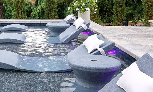 Submerged lounge chairs with white cushions and cup holders rest on a sun shelf under purple lights, perfect for in-pool use. The scene is enhanced by Ledge Loungers Signature Standard Side Table and a white flower arrangement among lush greenery.