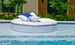 Relax in style on Ledge Loungers Signature Round Sunbed, a white circular inflatable float with cushioned seating, a central cup holder, and blue/white pillows. It sits beautifully on the water against a backdrop of bricks, lush greenery, and a nearby building.