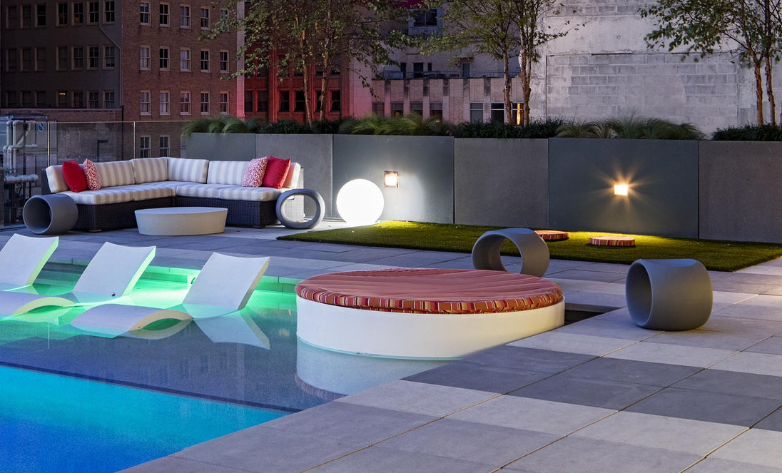 Relax on this modern rooftop terrace with a pool featuring submerged lounge chairs, a Ledge Lounger Signature Round Sunbed, striped sofa with red pillows, ambient lighting, and lush greenery. Enjoy the perfect cityscape backdrop for unwinding.