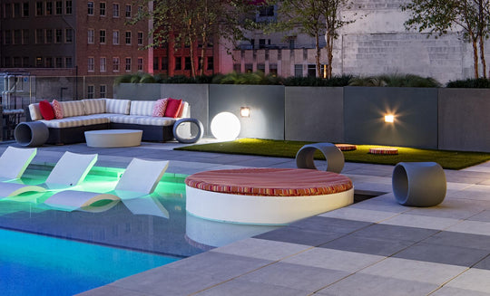 Relax on this modern rooftop terrace with a pool featuring submerged lounge chairs, a Ledge Lounger Signature Round Sunbed, striped sofa with red pillows, ambient lighting, and lush greenery. Enjoy the perfect cityscape backdrop for unwinding.